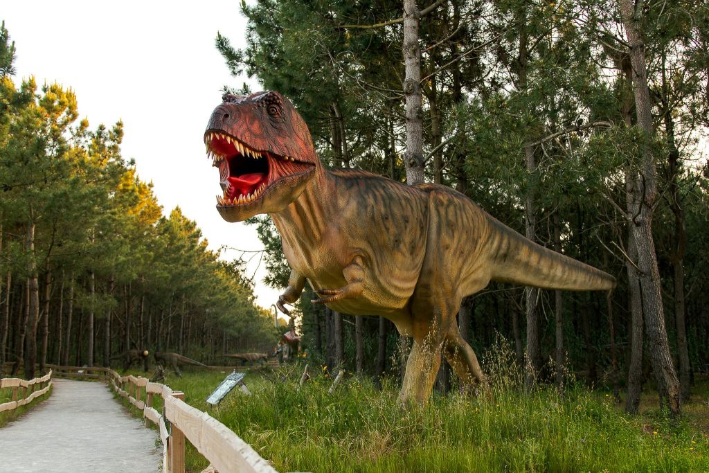 푸른 나무와 식물로 둘러싸인 커다랗고 입이 트인 T-Rex 동상이 있는 숲 속 통로.