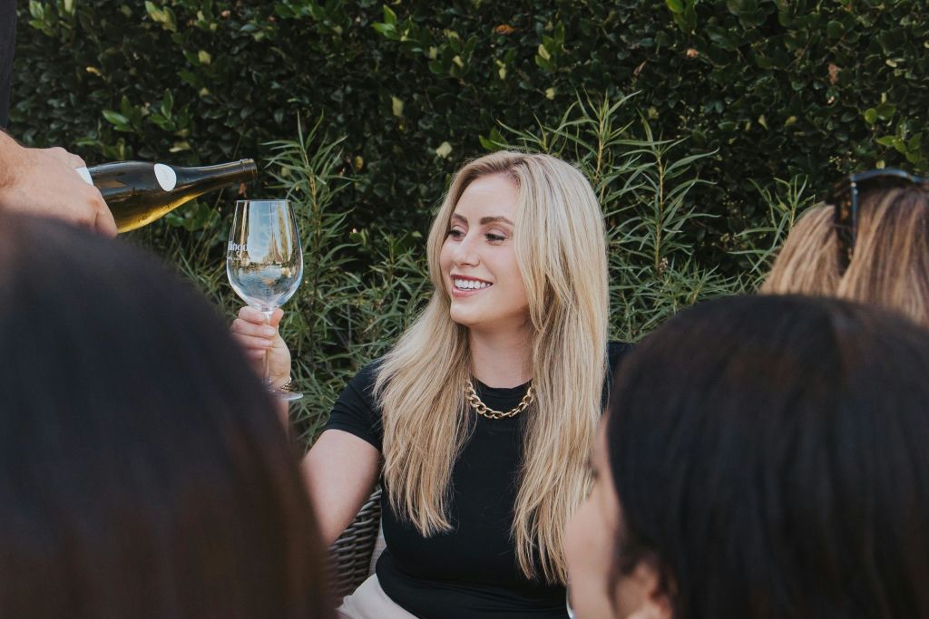 긴 금발 머리를 한 여성이 미소를 지으며 와인 잔을 들고 있습니다. 녹색 단풍이 배경입니다.