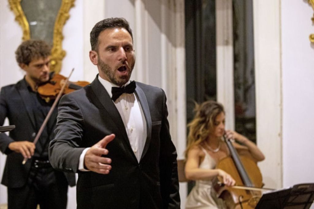 검은색 턱시도를 입은 남성이 실내에서 바이올리니스트와 첼리스트의 공연을 배경으로 열정적으로 노래합니다.