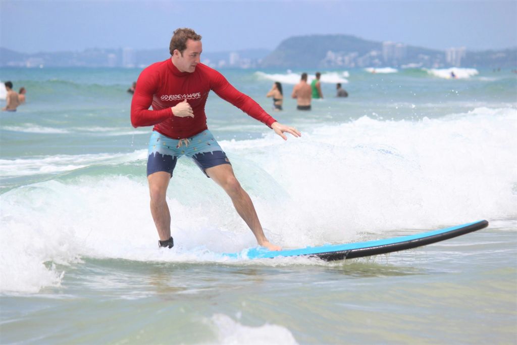 빨간색 셔츠와 파란색 반바지를 입고 해변에서 서핑하는 남성과 다른 사람들이 수영을 하고 있습니다.