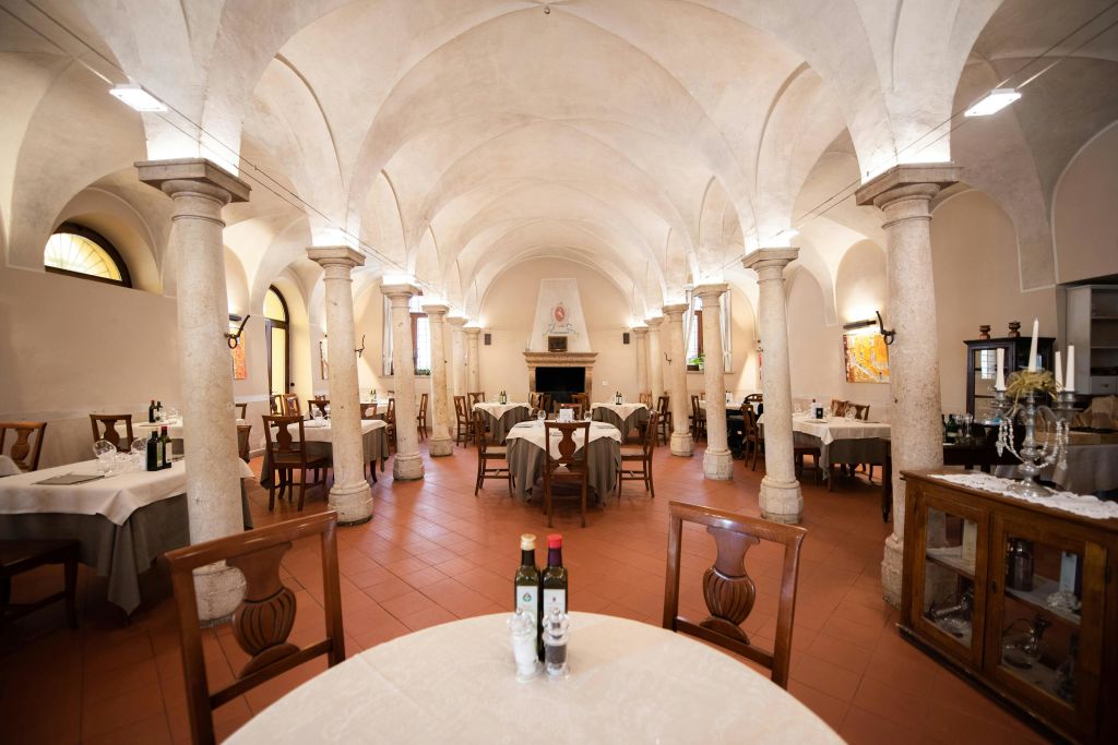 아치형 천장, 석조 기둥, 깔끔하게 정리된 테이블에 흰색 식탁보와 올리브 오일 병을 갖춘 우아한 레스토랑입니다.