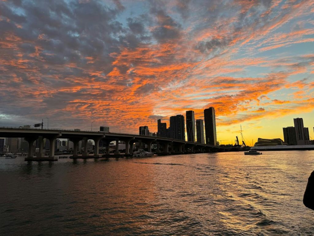 높은 건물과 물 위의 보트를 포함한 다채로운 하늘과 도시의 스카이라인이 있는 석양의 물 위 다리.
