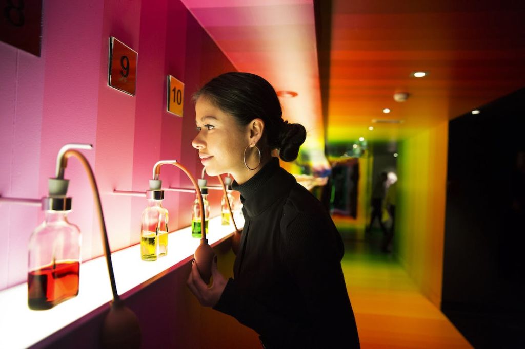 벽에 번호가 매겨진 패널이 있는 활기차고 다양한 색상의 방에서 후프 귀걸이를 한 여성이 유리병에서 냄새를 맡고 있습니다.