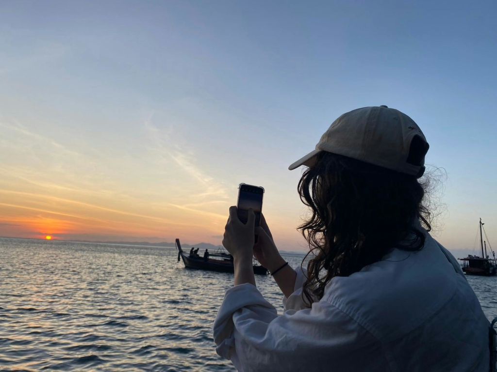 흰색 셔츠와 모자를 쓴 사람이 해질녘 바닷가에서 보트를 배경으로 휴대전화를 들고 사진을 찍고 있습니다.