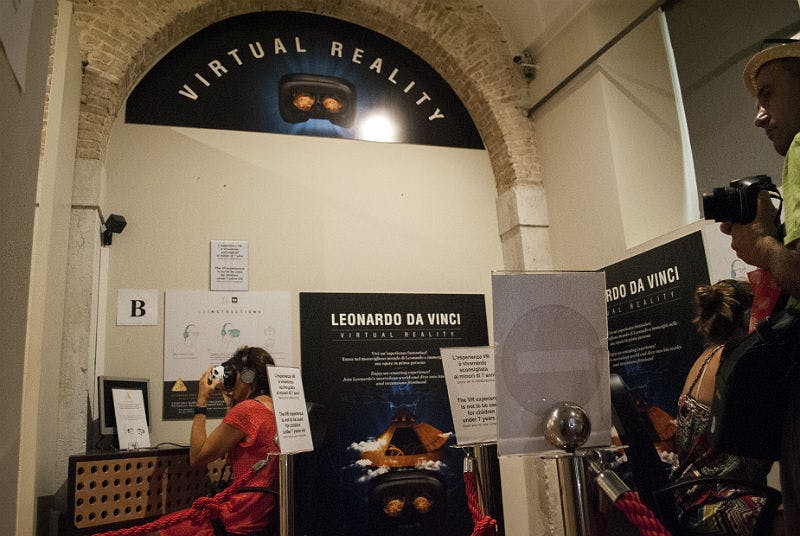 안내 포스터와 함께 'Leonardo da Vinci Virtual Reality' 라고 적힌 디스플레이 앞에서 VR 헤드셋을 사용하는 사람.