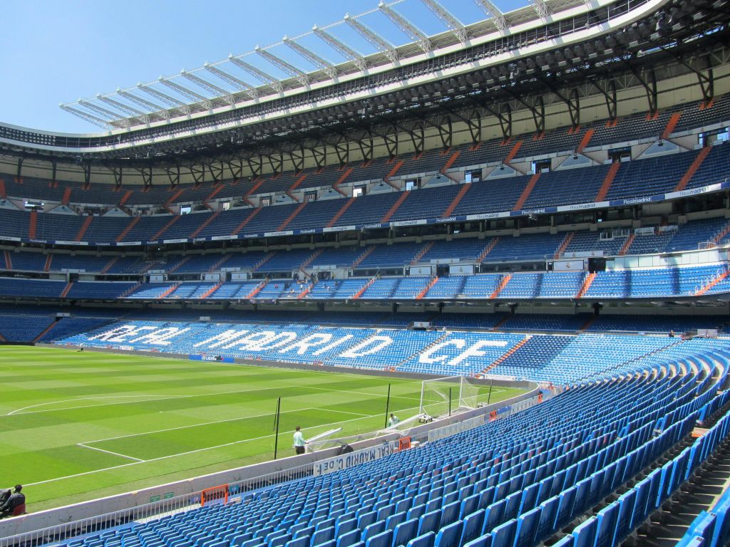 파란색 좌석과 "Real Madrid C.F." 레터링이 있는 빈 축구 경기장. 푸른 들판과 맑고 푸른 하늘.