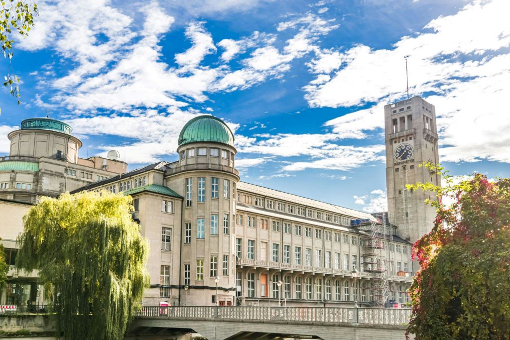 밝고 푸른 하늘 아래 녹색 돔과 시계탑이 있는 커다란 역사적인 건물로, 전면에 나무와 다리가 있습니다.