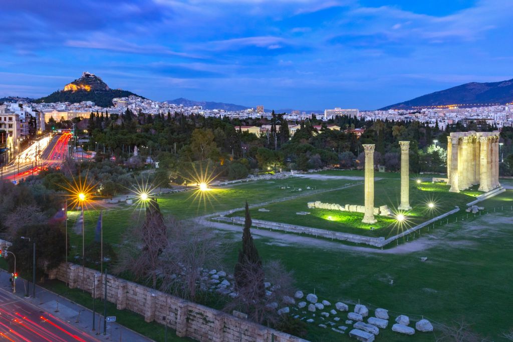 해질 무렵 도시와 언덕을 배경으로 녹음으로 둘러싸인 조명으로 빛나는 두 개의 기둥이 있는 고대 유적지.