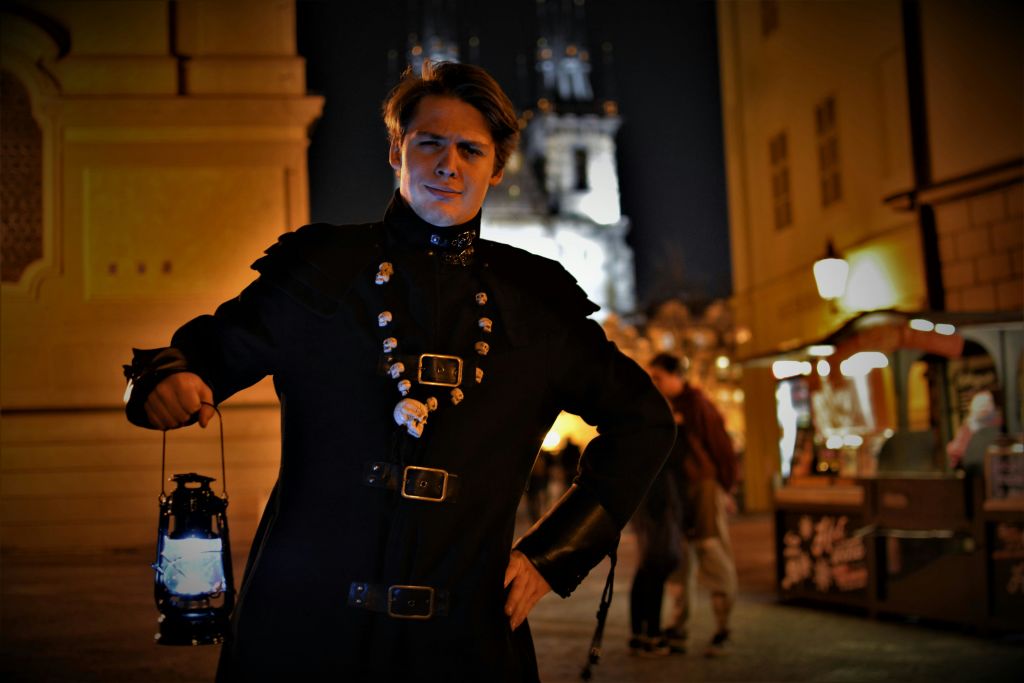 어두운 옷을 입은 사람이 등불을 들고 밤에 야외에 서 있습니다. 배경에는 조명이 켜진 건물과 다른 사람들이 보입니다.