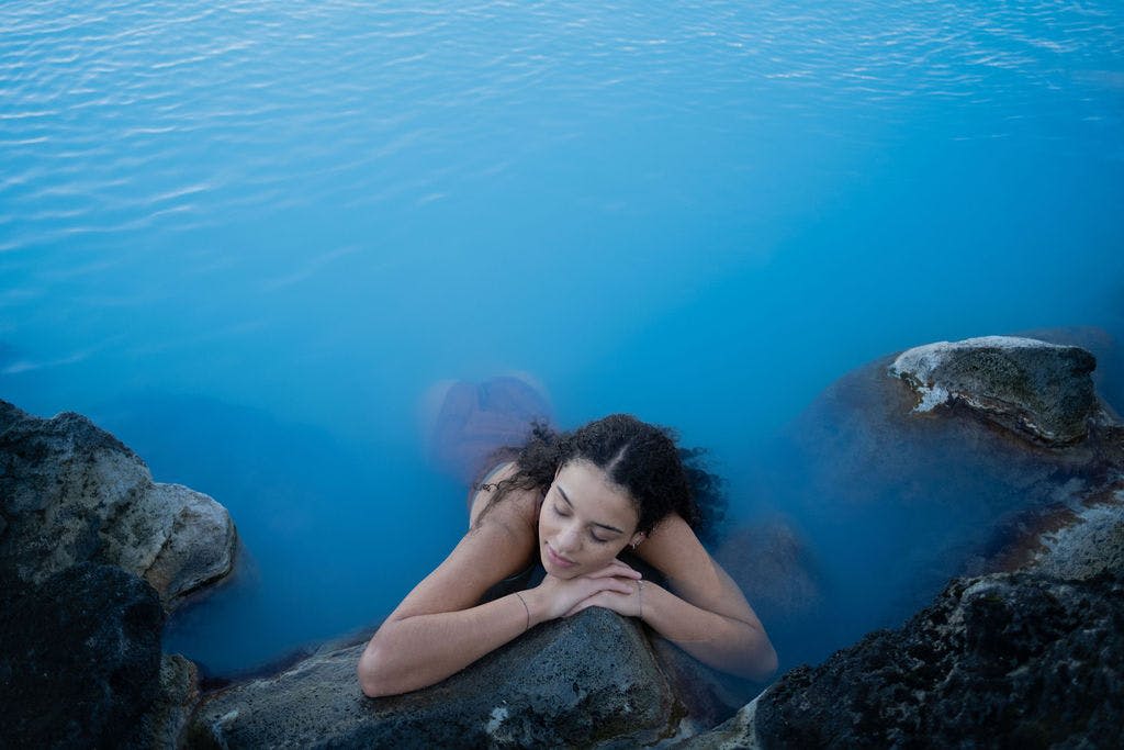한 사람이 물과 바위로 둘러싸인 눈을 감고 바위에 기대어 지열 푸른 수영장에서 휴식을 취하고 있습니다.