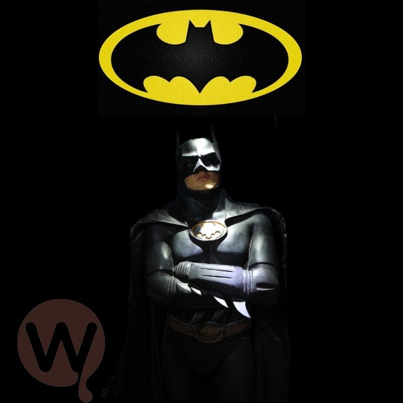 배트맨 의상을 입은 사람이 검은색 배경의 노란색 배트맨 로고 아래에 팔짱을 끼고 서 있습니다. 왼쪽 하단에 갈색 로고가 있습니다.