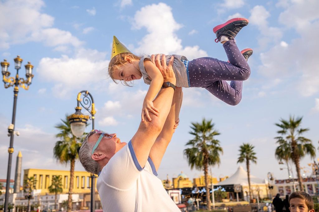 어른이 파티 모자를 쓴 웃는 아이를 공중으로 들어 올립니다. 야자수와 건물이 배경으로 보입니다.