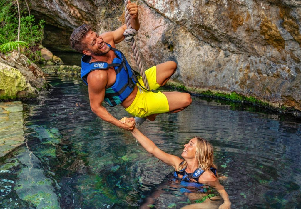 노란색 반바지를 입은 남성과 파란색 수영복을 입은 여성이 구명 조끼를 입고 미소를 짓고 바위 물 위에서 밧줄 그네를 타고 놀고 있습니다.