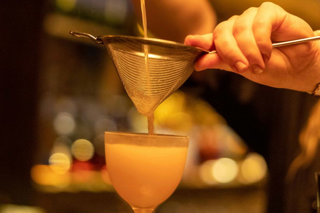 흐릿한 배경 조명이 있는 따뜻한 분위기의 유리잔에 스트레이너를 통해 칵테일을 붓습니다.