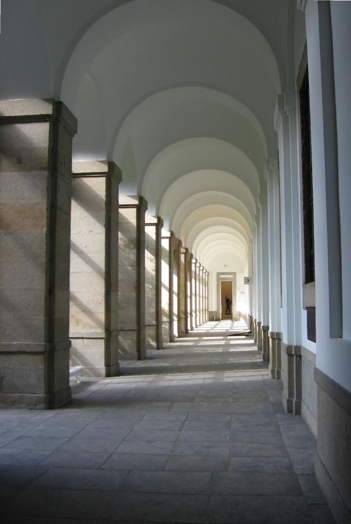 반복되는 기둥과 그림자가 있는 햇살이 비치는 석조 아치형 통로로, 한 사람이 맨 끝에 서 있는 먼 출입구로 이어집니다.
