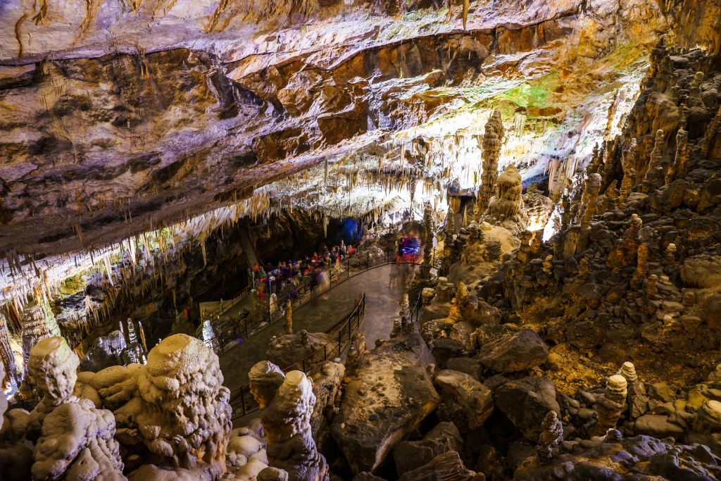 한 무리의 사람들이 종유석과 종유석이 있는 조명 동굴을 둘러보며 지정된 길을 따라 걷고 있습니다.