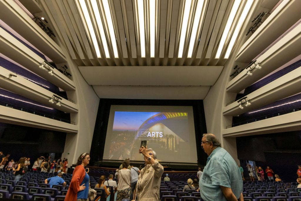 "LES ARTS" 라는 글자가 표시된 대형 스크린 앞에서 사진을 찍으며 층을 이루는 좌석이 있는 극장 내부에 있는 사람들.