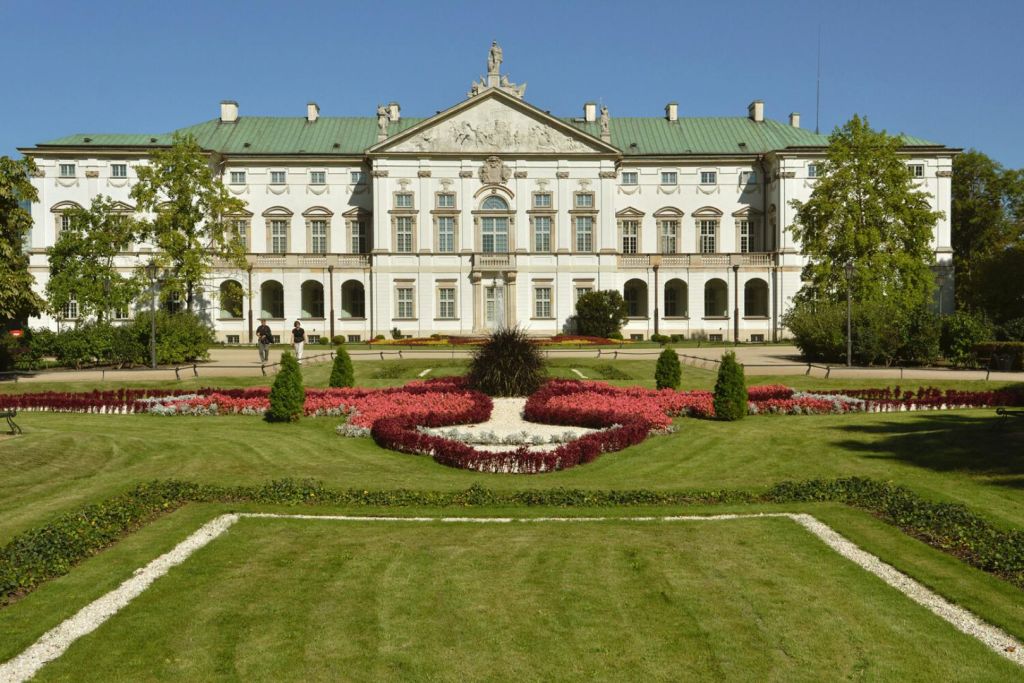 푸른 지붕이 있는 대형 신고전주의 건물, 화려한 외관, 붉은색과 흰색 꽃으로 단장한 정원, 맑고 푸른 하늘 아래.