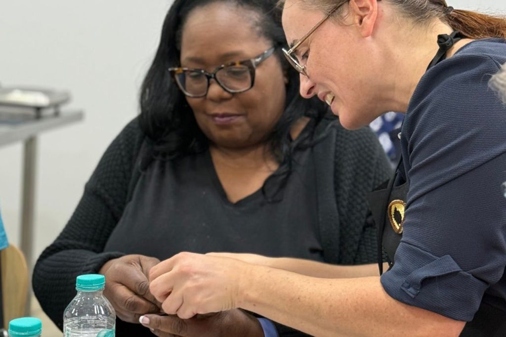 두 명의 여성이 테이블에서 상호 작용하고 있으며, 한 여성은 다른 여성이 작업을 수행하도록 돕고 있습니다. 테이블 위에 물병이 놓여 있습니다.