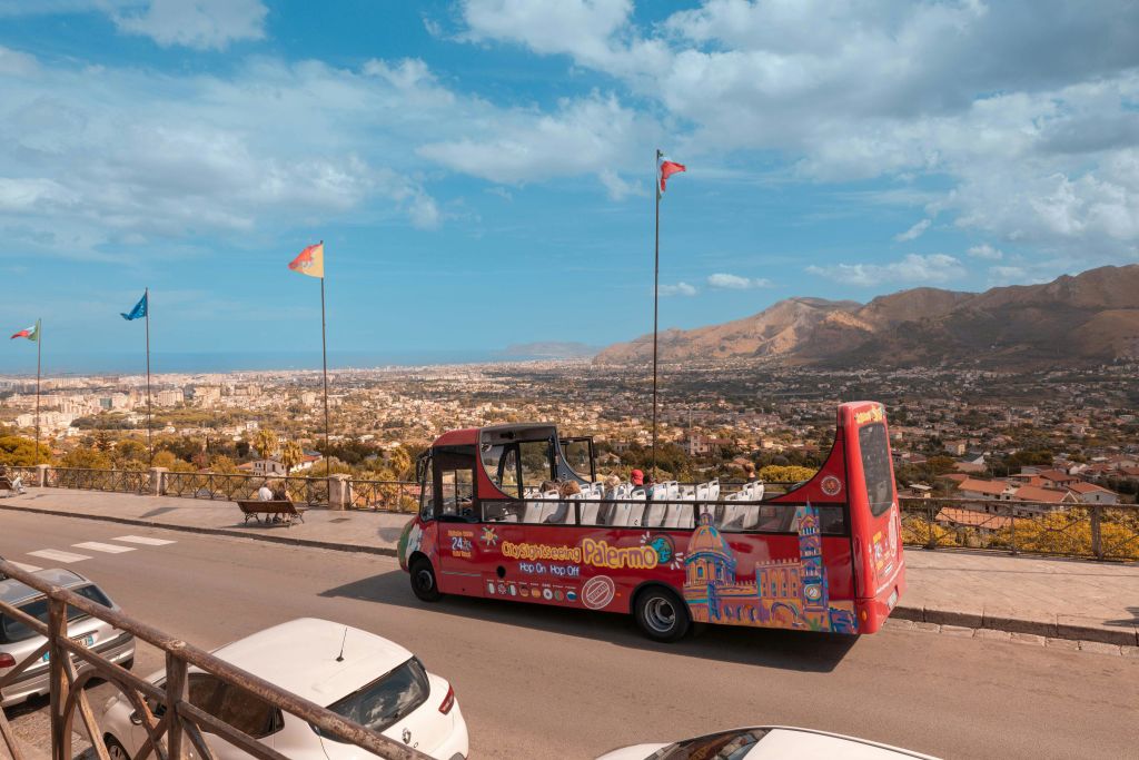 "팔레르모" 가 적힌 빨간색 오픈탑 투어 버스가 도시와 산이 내려다보이는 경치 좋은 전망대 근처에 주차되어 있습니다.