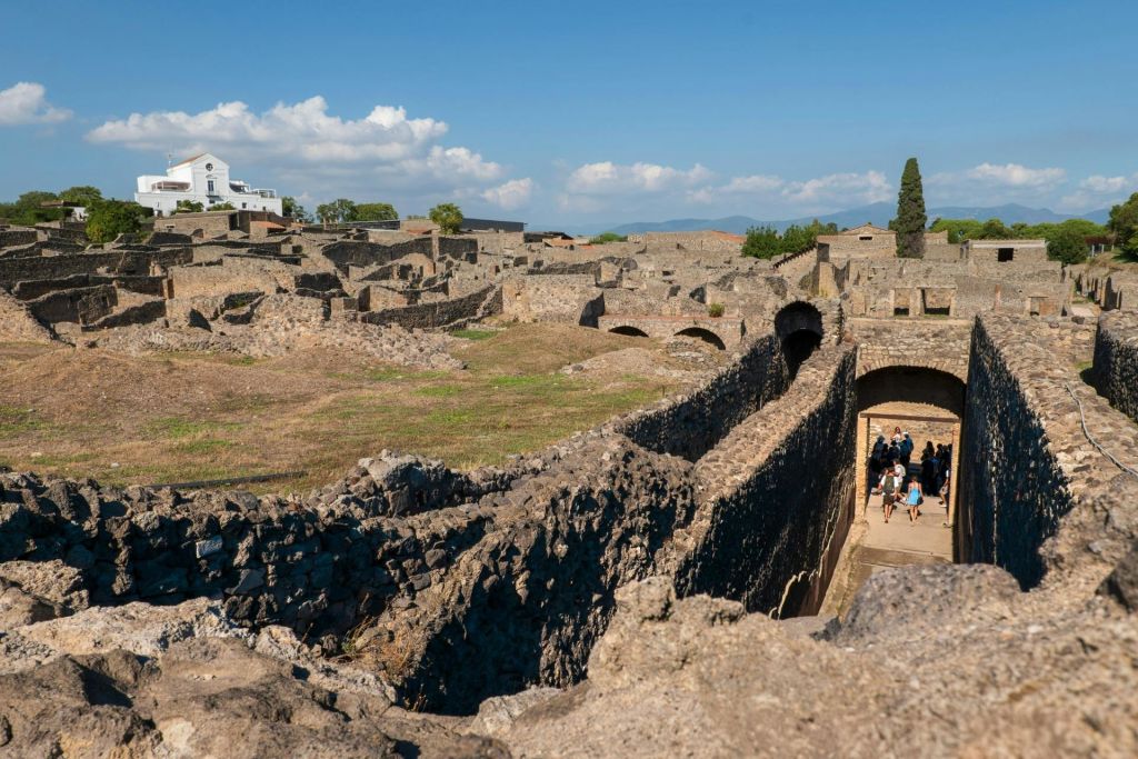 맑고 푸른 하늘 아래 돌담과 통로, 아치가 있는 고대 유적지. 방문자 그룹이 사이트를 둘러봅니다.