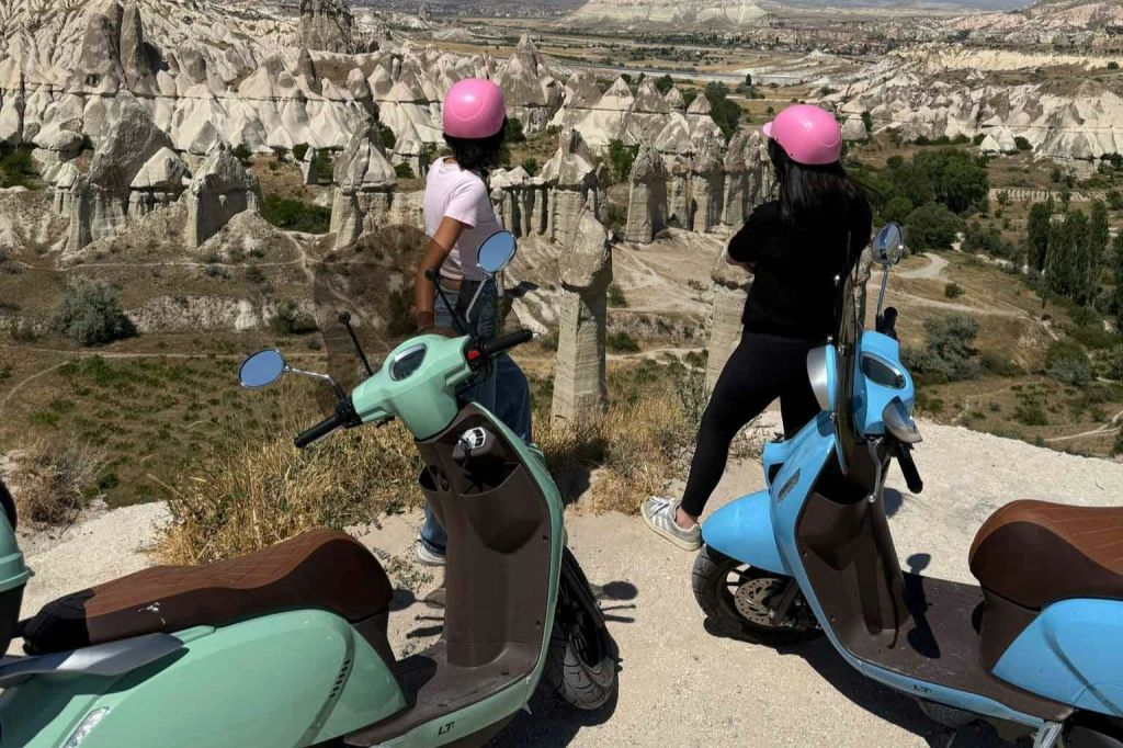 분홍색 헬멧을 쓴 두 명의 여성이 두 대의 스쿠터 옆에 서서 험준한 풍경의 암석 지대가 내려다보입니다.