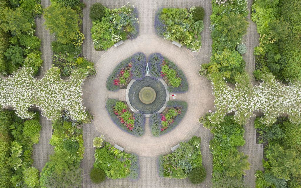 대칭형 화단이 있는 통로, 벤치, 울창한 녹지로 둘러싸인 원형 정원 분수의 공중 전망.