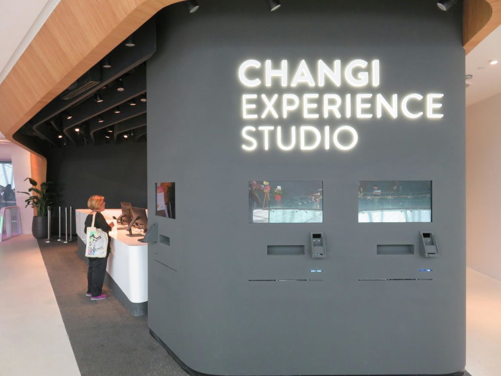 한 여성이 창이 체험 스튜디오의 흰색 카운터에 서 있는데, 안내 키오스크와 커다란 조명 간판이 보입니다.