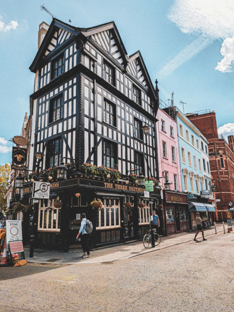 "쓰리 그레이하운드 (The Three Greyhounds)" 라는 이름의 코너 펍으로, 반목으로 지어진 건축물, 화려한 색상의 건물이 인접해 있으며 거리에 보행자가 있습니다.