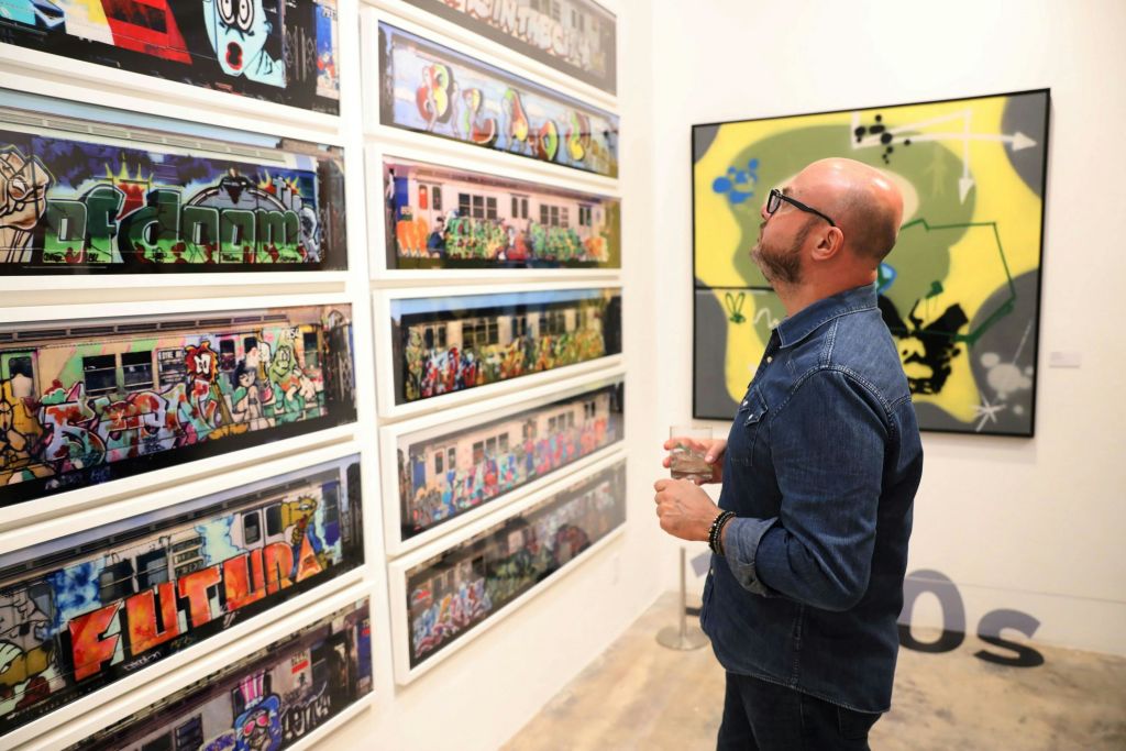 데님 셔츠를 입은 한 남성이 음료를 들고 갤러리에서 액자에 담긴 그래피티 아트를 살펴보고 있습니다.