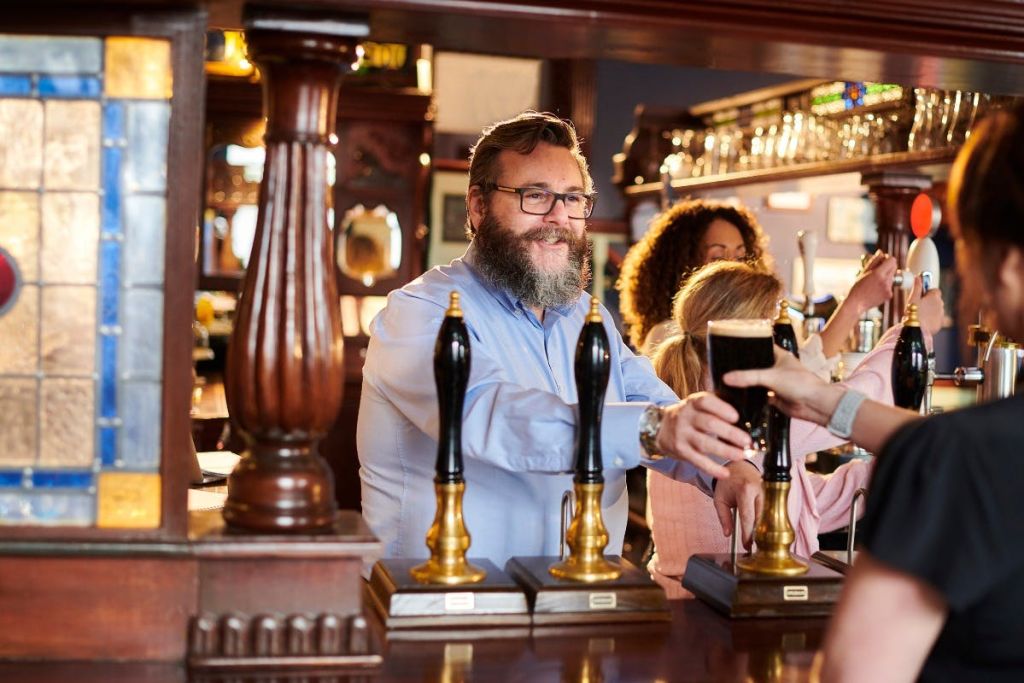 수염을 기른 바텐더가 전통적인 펍에서 손님에게 음료를 제공합니다. 나무 장식과 탭이 보입니다.