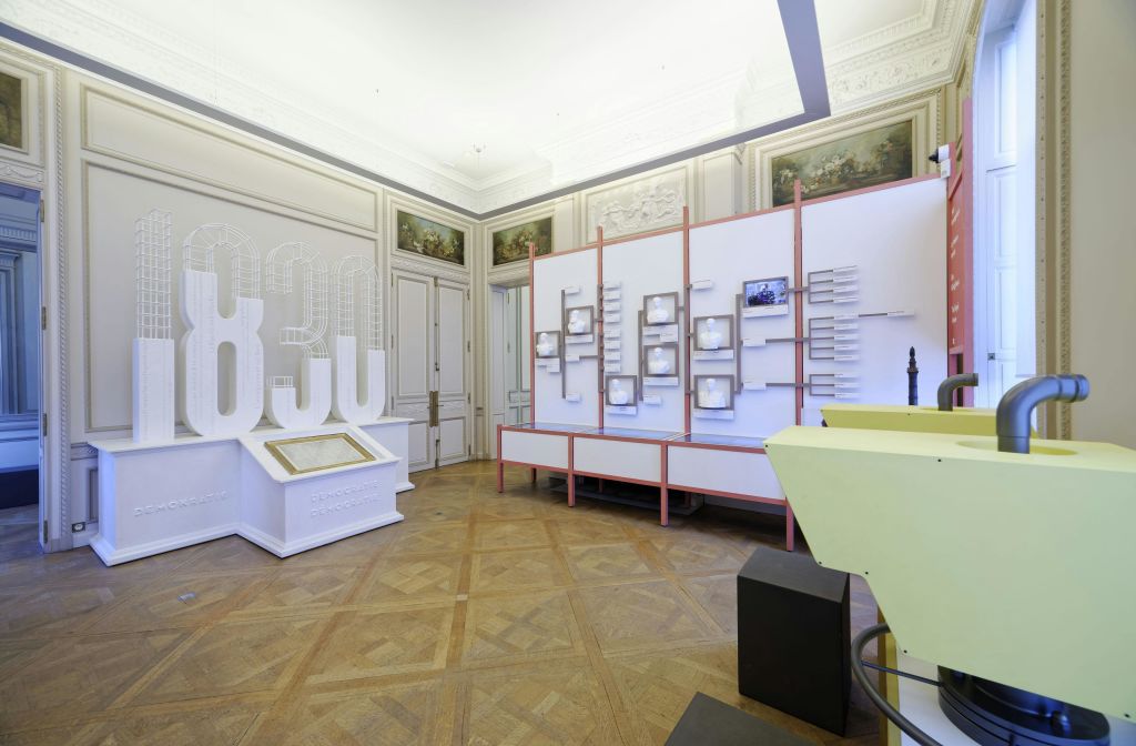 흰색과 분홍색의 전시 구조물, 액자에 담긴 벽면 예술 작품, 나무 바닥에 있는 터치스크린 단상이 있는 현대적인 전시실입니다.