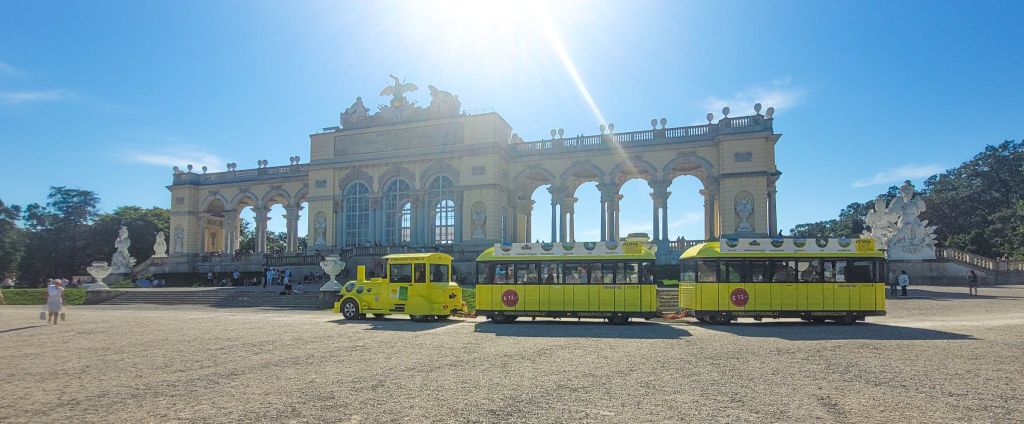 밝고 화창한 하늘 아래 아치와 조각상이 있는 웅장하고 유서 깊은 건물 앞에 노란색 관광 열차가 있습니다.