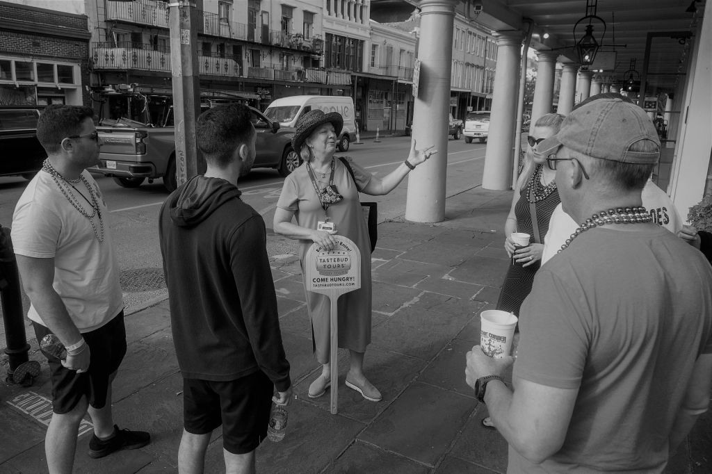 모자를 쓴 한 여성이 근처에 기둥과 건물이 있는 도심의 보도에 서서 투어를 진행합니다.