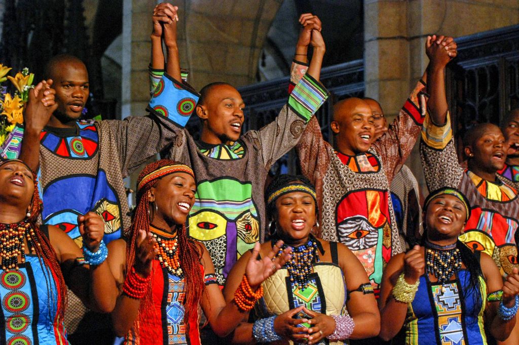 다채로운 전통 복장을 한 사람들이 석조 아치가 있는 활기찬 실내 환경에서 노래를 부르고 손을 잡고 있습니다.