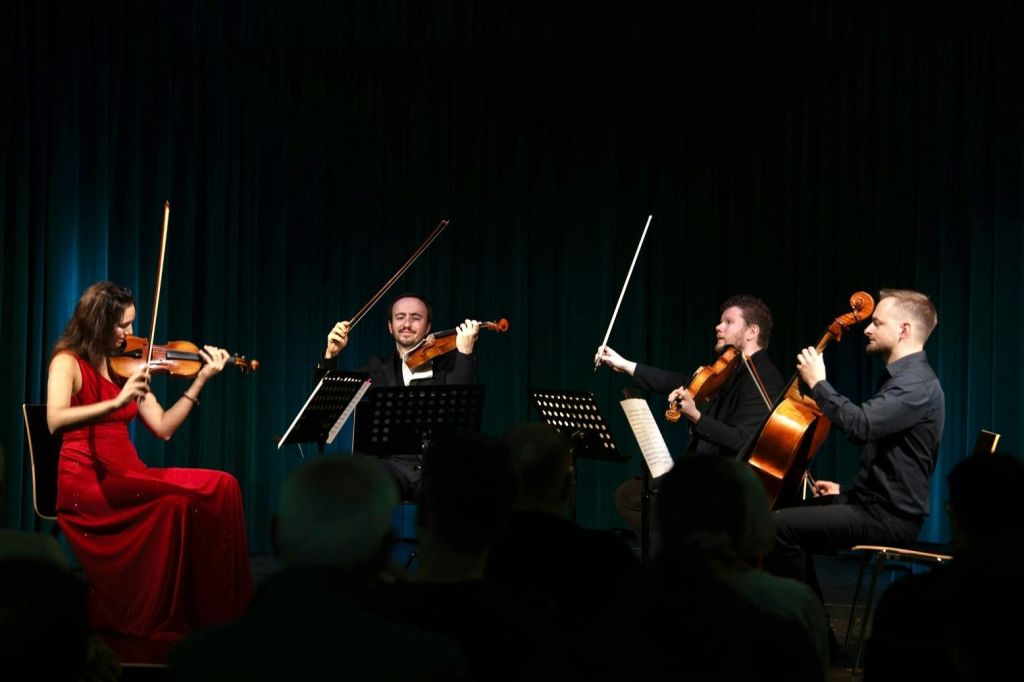 세 명의 뮤지션이 무대에서 공연하며, 각자 바이올린을 연주합니다. 여성은 빨간색 드레스를 입고, 두 남성은 정장 차림을 하고 있습니다.