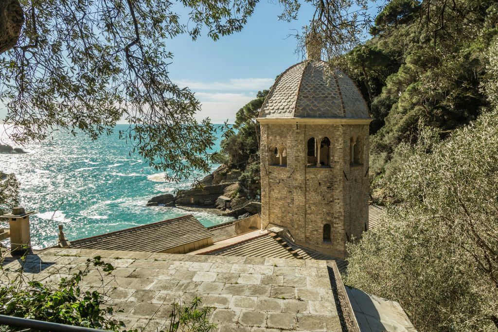 나무와 단풍으로 둘러싸인 바위 해안선과 청록색 바다가 내려다보이는 돔 지붕의 석조 건물.