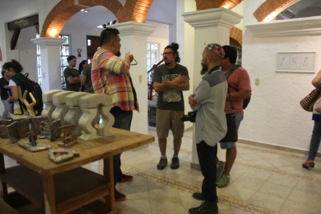 흰색 벽과 벽돌 아치가 있는 방에 서 있는 네 명의 남성이 돌조각이 놓인 나무 테이블 근처에서 대화를 나누고 있습니다.