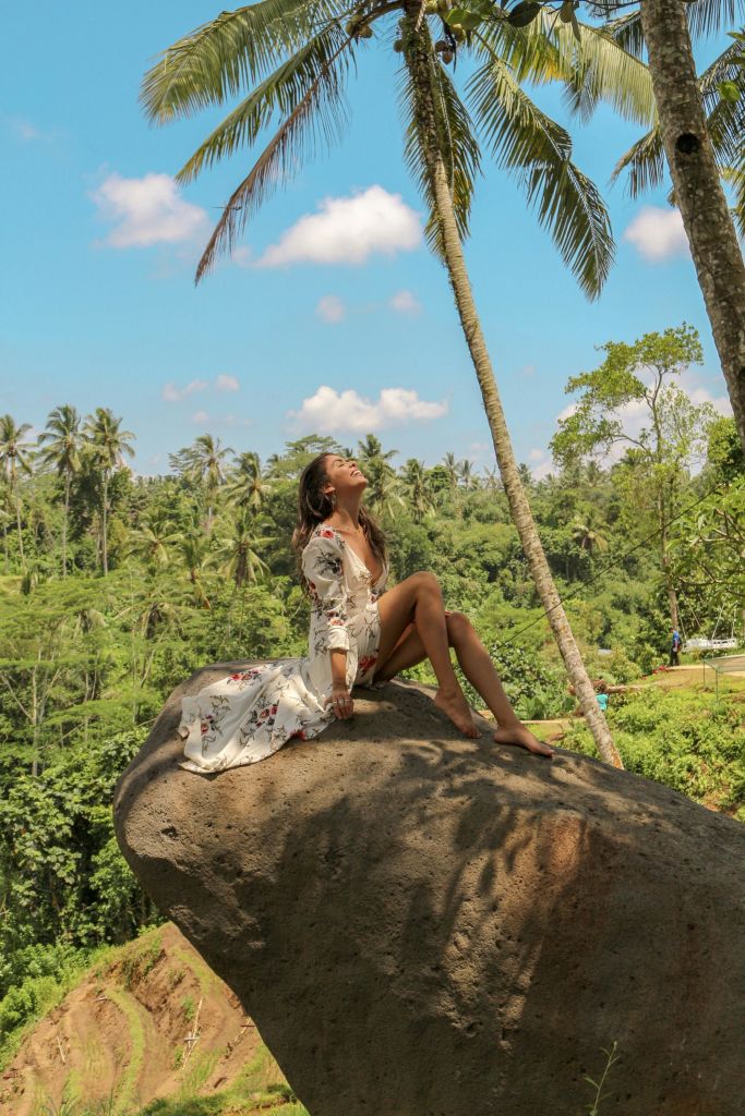 부분적으로 흐린 하늘 아래 푸른 녹음과 높은 야자수로 둘러싸인 커다란 바위에 꽃무늬 드레스를 입은 한 여성이 앉아 있습니다.