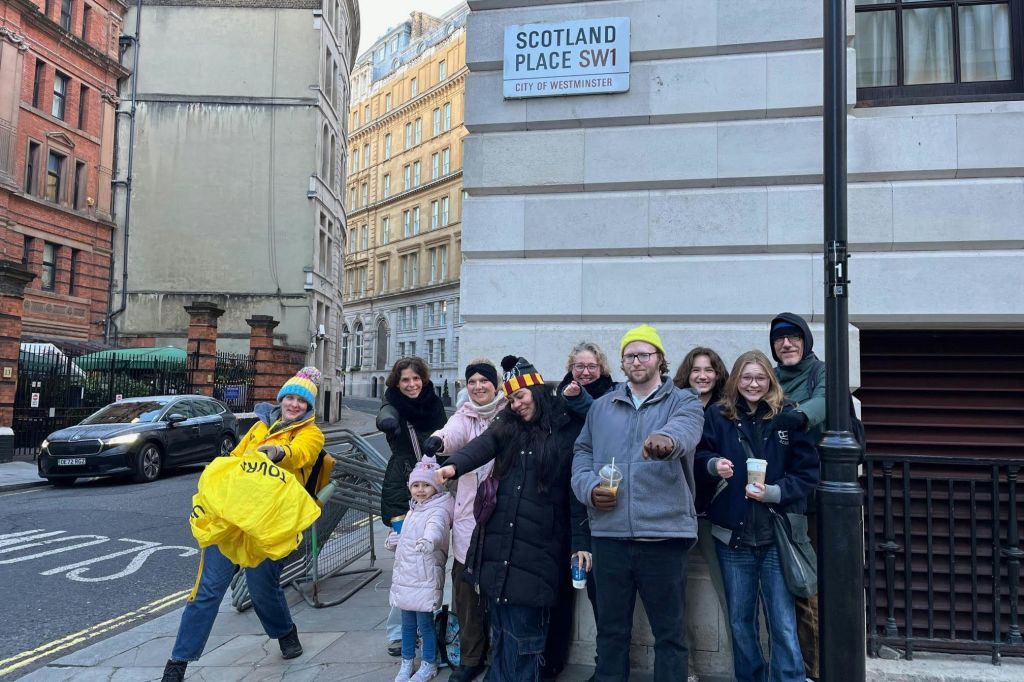 한 무리의 사람들이 건물 근처의 도시 거리에서 "Scotland Place SW1, City of Westminster" 라는 표지판이 있는 사진을 찍기 위해 포즈를 취하고 있습니다.