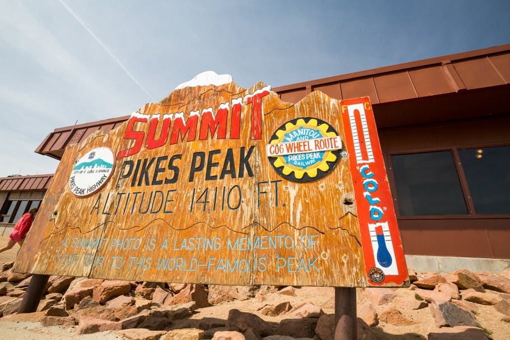 파이크스 피크 서밋 (Pikes Peak Summit) 의 풍화된 나무 표지판은 고도를 자랑하며, 바위와 건물을 배경으로 둘러싸여 있습니다.