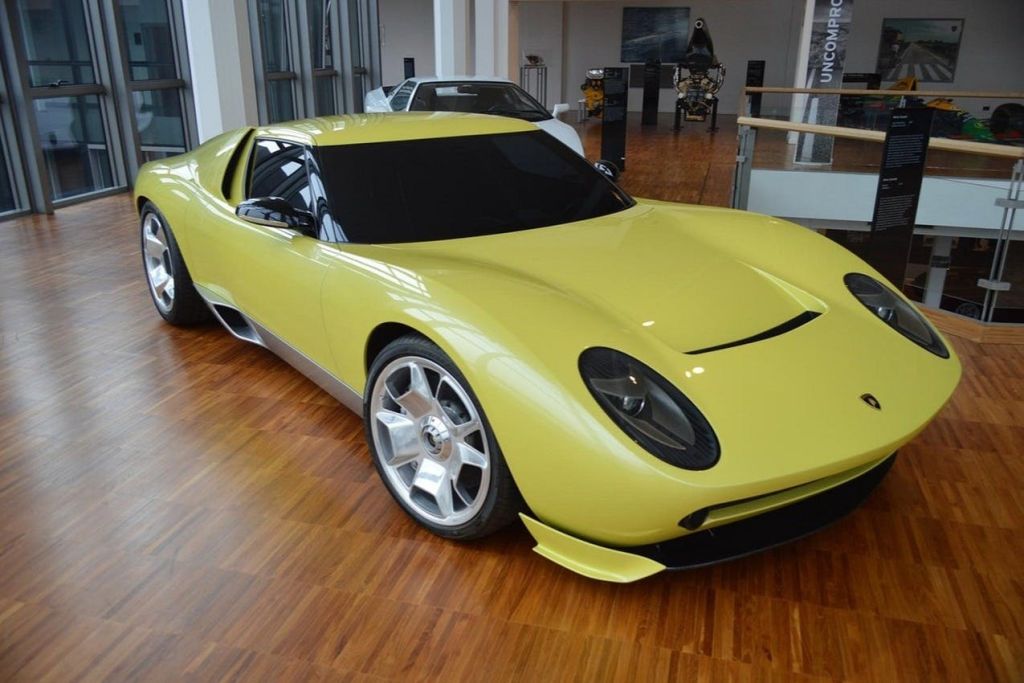 나무 바닥이 있는 쇼룸에 노란색 스포츠카가 전시되어 있습니다. 배경에는 또 다른 자동차와 전시품이 보입니다.