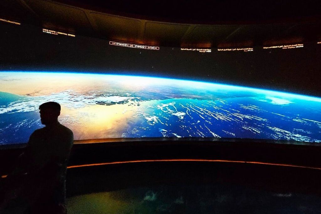 어두운 방의 대형 스크린에 표시된 공간에서 바라본 지구의 모습, 왼쪽에 사람의 실루엣의 일부가 보입니다.