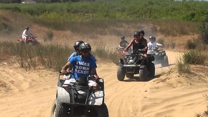 몇몇 사람들은 헬멧과 캐주얼한 옷을 입고 흙길에서 ATV를 타고 다닙니다. 잔디와 수풀이 배경으로 보입니다.