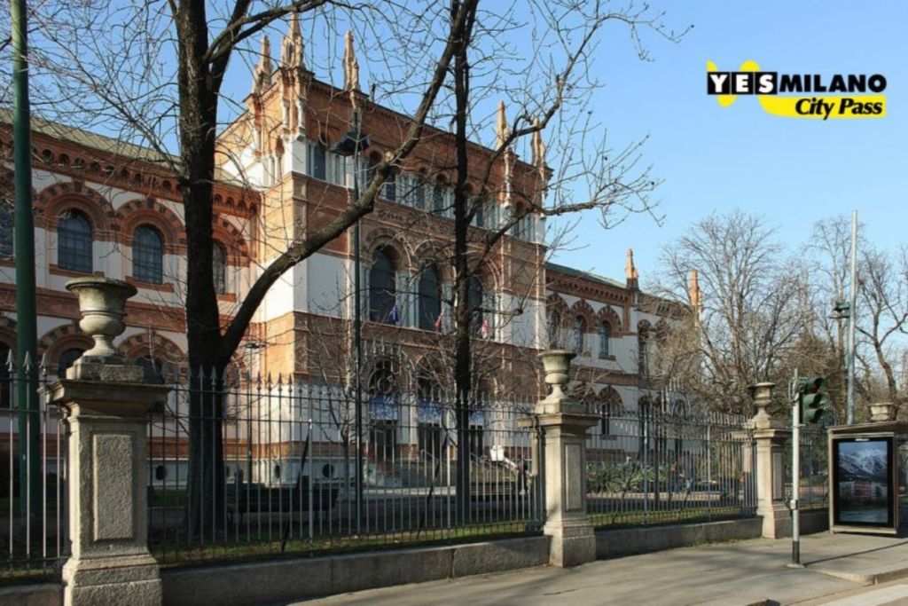 검은 철제 울타리와 맨 나무로 둘러싸인 아치형 창문이 있는 유서 깊은 벽돌 건물입니다.