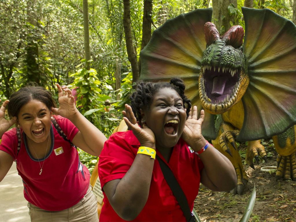 울창하고 녹음이 우거진 야외 환경에서 두 소녀가 웃으며 공룡 상을 흉내냅니다.