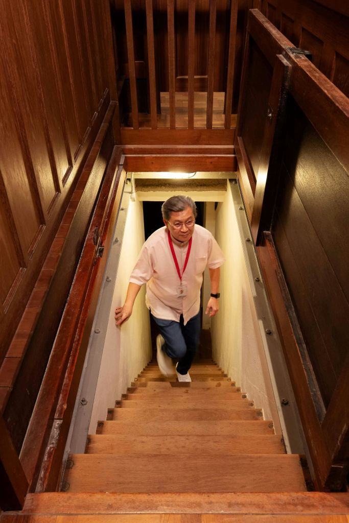 흰색 셔츠와 빨간 방아끈을 두른 남성이 조명이 어두운 지하실에서 좁은 나무 계단으로 올라갑니다.