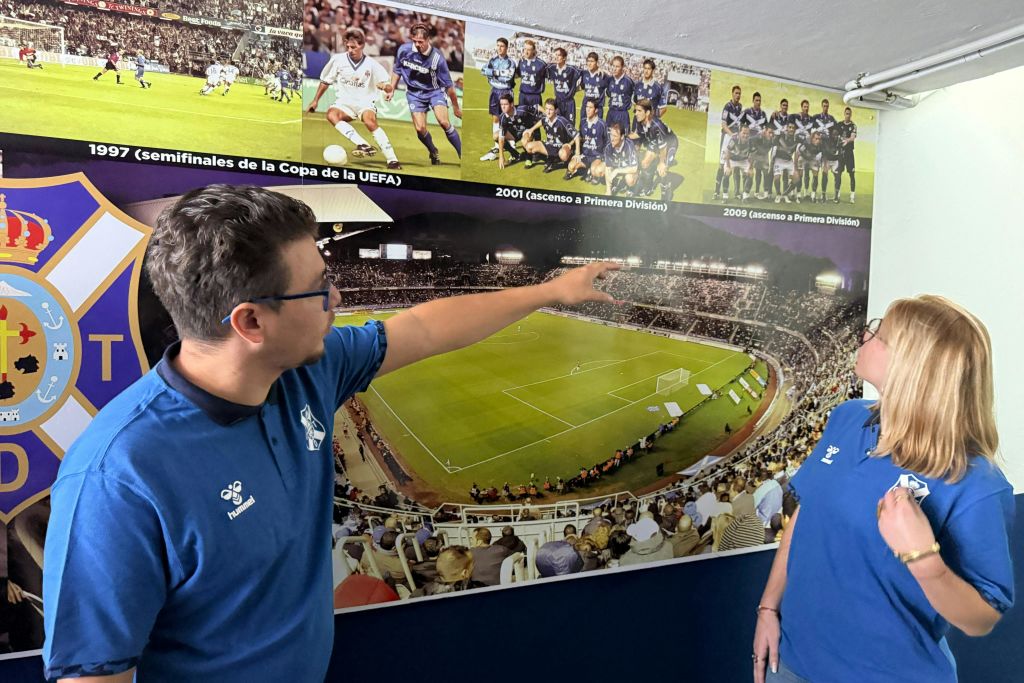 파란색 셔츠를 입은 한 남성이 근처에 커다란 경기장 이미지와 팀 로고가 있는 벽에 걸린 유서 깊은 축구 경기 사진을 가리키고 있습니다.