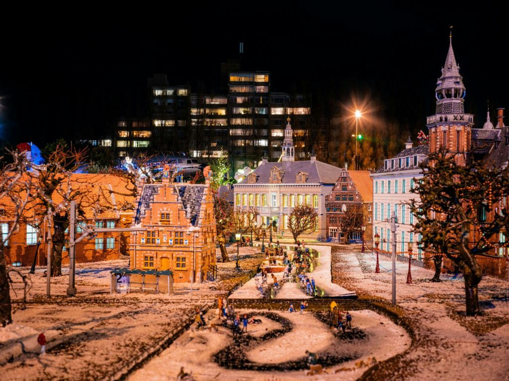 야간에는 도시의 불빛을 배경으로 조명이 켜진 건물, 나무, 통로에 작은 인물이 있는 눈 덮인 미니어처 마을 장면이 펼쳐집니다.