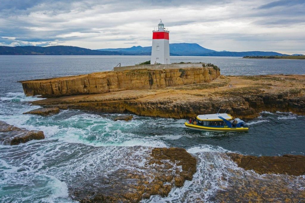 보트는 흐린 하늘 아래 흰색과 빨간색 등대를 배경으로 바위 해안 근처를 항해합니다.